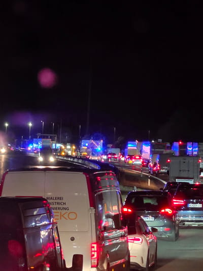 The image depicts a traffic jam at night on a highway, possibly a German Autobahn, with numerous vehicles including cars and trucks. Emergency lights, primarily blue and red, are visible on some vehicles in the distance, suggesting an incident or road closure. The road surface appears wet, indicating recent rain. In the foreground, a white van with text "MALEREI NSTRICH NIERUNG EKO" is visible. Several cars with their brake lights illuminated are also present. The sky is dark, with some distant streetlights and vehicle headlights providing illumination. There is a pinkish, blurry light artifact in the upper left quadrant of the image. No people are clearly discernible in the foreground or midground. The scene conveys a sense of disruption and congestion on a major roadway. The mention of Uettingen, Germany, implies this traffic jam is occurring within or near that locality.