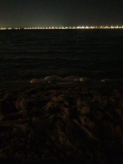 This night photograph captures a tranquil coastal scene in Al `Aziziyah, Saudi Arabia, looking out over a body of water, likely the Persian Gulf. The image is composed of three distinct horizontal sections.

In the foreground, the dark, textured sand of a beach occupies the lower portion of the frame, showing subtle undulations and patterns. Just beyond the sand, at the water's edge, faint, lighter patches of what appear to be foam or gentle surf are visible, indicating small waves breaking softly on the shore.

The middle ground is dominated by the dark expanse of water, which stretches across the majority of the image. Its surface shows subtle ripples, reflecting the calm conditions.

Across the distant horizon, a long, continuous line of bright artificial lights defines the boundary between the dark water and the dark night sky. These lights, varying from warm yellow-orange to white, are numerous and evenly spaced, suggesting a distant city, town, or industrial area lining the far shore. Their glow creates a stark, luminous contrast against the otherwise dark environment.

The time of day is clearly night, with no natural light sources like the sun or moon visible, highlighting the artificial illumination. No people or specific activities are depicted, giving the scene a quiet and contemplative atmosphere. The weather appears clear, allowing the distant lights to be seen distinctly. No visible text is present in the image.