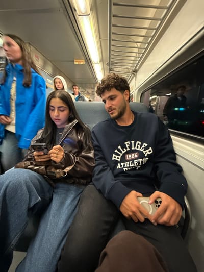 The image displays the interior of a passenger train car, located in East Rutherford, United States. The scene is brightly illuminated by overhead fluorescent lighting. In the foreground, a female with long, dark, straight hair, wearing a dark brown leather-like jacket and blue wide-leg jeans, is seated on the left. She is engaged with a dark-cased smartphone held in both hands, displaying several rings on her fingers. A brown shoulder bag with a gold-colored buckle rests on her lap. To her immediate right, a male with dark curly hair and light facial stubble is seated, wearing a navy blue crewneck sweatshirt with "HILFIGER 1985 ATHLETIC DEPT." printed on the chest. He holds a light-colored smartphone on his lap and is looking towards the female.

In the mid-ground, a female with long dark hair, wearing a bright blue hooded jacket, is partially visible on the far left, appearing to move away from the camera. Further down the aisle, other passengers are visible, including a female in a light-colored hooded top and a male with light hair wearing a dark jacket. The train interior features gray upholstered seating, white ceiling panels, and metal overhead luggage racks on the right side. A red and yellow "EMERGENCY EXIT" sign with an arrow is mounted on the ceiling above the aisle. Darkened windows on the right side of the train reflect interior lights and blurred exterior elements.