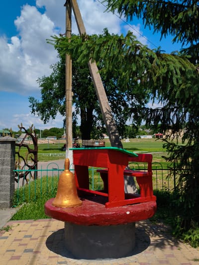 The image captures an outdoor scene in Velyka Bahachka, Ukraine, featuring a decorative red school desk and bell structure situated on a circular, red-painted base atop a concrete cylindrical foundation. The structure is adorned with a green accent along the desk's edge and a golden bell with a clapper is placed prominently on the red base. Behind the desk, a large concrete pole supports a leaning, angled concrete support beam, partially obscured by the lush green branches of an adjacent evergreen tree. A metal fence with vertical green bars encloses the area in front of the structure, and beyond the fence lies a grassy field. A decorative metal gate with a branch motif is visible to the left, next to a grey stone pillar. In the background, trees and buildings can be seen, and a red tractor is parked in the field. The sky above is blue with scattered white clouds. The ground in the foreground is paved with a pattern of yellow and reddish-brown interlocking bricks.