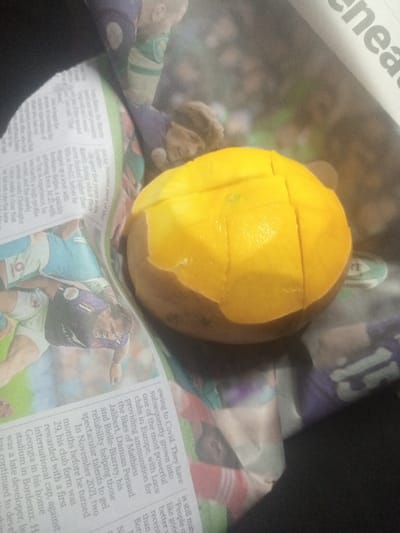A sliced mango sits on a newspaper. The skin is visible along the bottom half. The mango's flesh is cut into a grid pattern, ready to be eaten. The newspaper is printed with a collage of sports images. Text is visible, including words such as "Covid," "Europe," "talents," "Bordeaux," "international," and "clubs." A portion of a person's dark clothing is visible on the side.