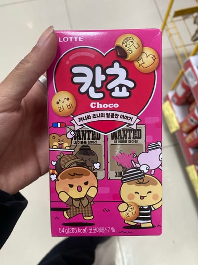 A person is holding a box of Lotte Kancho Choco cookies. The box is pink with cartoon-like illustrations on it. The text on the box is in Korean. The brand name "LOTTE" is printed vertically on the left side of the box. Some cans are visible in the blurred background.