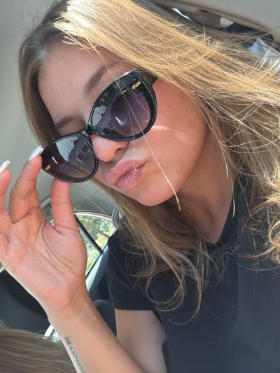 The image shows a close-up selfie of a young woman with long, blonde hair, wearing black sunglasses and a black t-shirt. She is holding the sunglasses with her left hand, which has white-painted fingernails. Her lips are slightly parted, giving a pouting expression. The background suggests she is in a car, with a glimpse of trees and sky visible through a window. A tattoo is visible on her left wrist. The scene is lit by natural sunlight, casting shadows on her face. The image is a selfie, taken from a high angle. There is no identifiable information to place this image in Palje Brdo, Croatia.