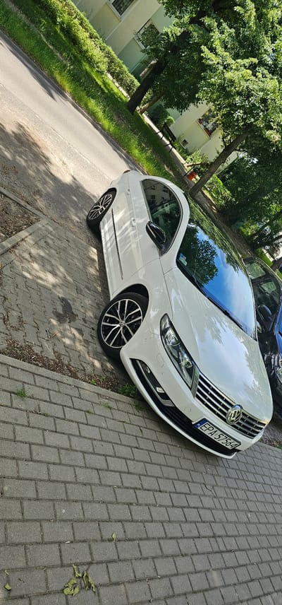 The image shows a white Volkswagen CC parked on a brick-paved sidewalk next to a road. The car has a black roof and tinted windows. The license plate is visible and reads "EPA 57564". The car is parked alongside a green grassy area with trees and a building in the background. The building appears to be a residential block with windows and some greenery on its facade. The trees are lush and full of leaves, suggesting it is daytime and likely a warm season. Sunlight casts shadows on the sidewalk and road, indicating a sunny day. The scene is set in Pabianice, Poland. There are no people visible in the image. The overall impression is a quiet, everyday street scene.