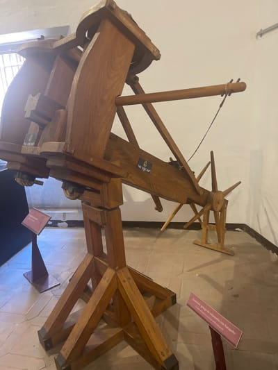 The image shows a wooden replica of an ancient catapult, likely a model designed for educational purposes. The catapult is constructed from thick wooden beams and has a complex system of levers and a projectile arm. Two informational plaques are visible in the foreground. One plaque, in Spanish and English, reads "Guerrero griego de infantería con Katapelte" and "Greek infantry warrior with catapult." The other plaque, partially visible, appears to be a description of the artifact itself. The background suggests an indoor setting, possibly a museum or exhibition space, with a tiled floor and a white wall. The lighting is somewhat dim, casting shadows on the floor and the artifact. The overall impression is of a historical exhibit. The information about the location in Toledo, Spain, is not present in the visual data and cannot be inferred.