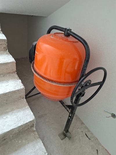 An orange, portable cement mixer with a black metal frame and steering wheel stands on a concrete floor in an unfinished interior space, likely under construction or renovation, located in Krupnik, Bulgaria. To the left, a rough concrete staircase ascends. The walls are light gray, consistent with recently applied plaster or paint. On the right-hand wall, a circular opening exposes blue, yellow/green, and black electrical wires. The concrete floor is dusty, with small pieces of debris present. No individuals are depicted in the scene.