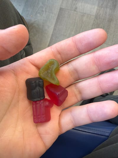 A close-up, medium shot depicts a person's open palm holding four assorted gummy candies. The candies are of various shapes and colors: a green heart shape, a red cube shape, a black rectangular block shape, and a red rectangular shape with indented lines. The person's skin tone appears fair. In the background, there are sections of gray flooring, a dark green fabric, and a blue, possibly leather, seat. The image gives no clues about the city or country.