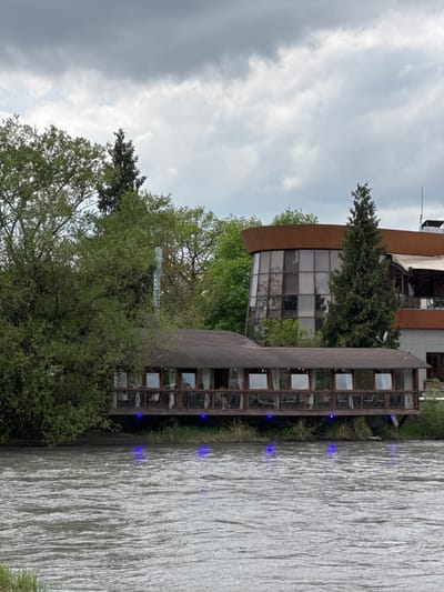 This image shows a multi-level building complex on the edge of a body of water in Zamankul, Russia. The architecture incorporates large windows and a cylindrical design element, partially obscured by trees. The sky is overcast with gray clouds. Ripples are visible on the surface of the water, and blue lights illuminate the side of the building closest to the water.