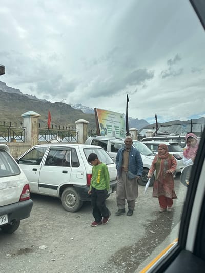 In Dras, India, under a cloudy sky, a group of people walk on a gravelly, wet road. The group consists of a young boy in a green long-sleeved shirt and dark pants, followed by an elderly man in a blue jacket and brown pants, and two women in colorful traditional clothing. One woman holds a white umbrella. Several cars are parked or stopped on the road, including a white Maruti 800. In the background, a fence with decorative pillars stands before a mountain range, with a red flag visible on a pole. A large banner with text and images is also present. The scene suggests a stop along a roadside in a mountainous region.