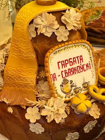 The image shows a close-up of a decorative loaf of bread in what appears to be a market or festival setting. The bread is elaborately decorated with edible elements that resemble fabric, flowers, and a traditional samovar with tea cups. A sign with Cyrillic script, "ГАРБАТА - СВАЯКОЎСКІ", is affixed to the bread, likely indicating its name or a related offering. The overall impression is one of a festive, culturally significant food display. The specific location in Вяйнянскі сельскі Савет, Belarus is not visually discernible from the image itself.