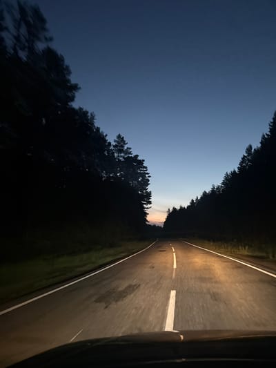 The image depicts a view from the perspective of a driver traveling on a two-lane asphalt road at dusk or night. The road has white lane markings. On either side of the road, there are dense, dark forests with tall coniferous trees. The sky above is a deep blue, transitioning to a lighter hue with a hint of orange near the horizon, suggesting the sun has recently set or is about to rise. The headlights of the vehicle illuminate the road ahead, casting a warm glow. The scene appears to be a rural or natural environment, likely a forested area. No people or specific landmarks indicating the city of Balak, Russia, are visible.