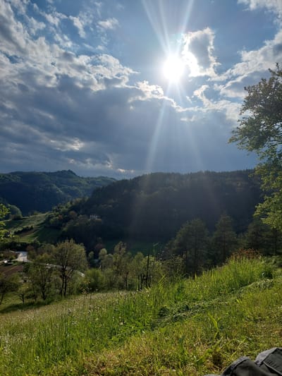 The image depicts a sunlit mountain landscape, likely captured in Kozarka, Bulgaria, due to its mountainous terrain. The foreground features a green meadow with tall grasses and scattered wildflowers, possibly marguerites. In the distance, a densely forested mountain ascends towards the sky, its slopes covered in coniferous and deciduous trees. At the base of the mountain, a winding road is visible with a few buildings. Above, the sky is partially cloudy with the sun shining brightly, casting rays downwards. A small portion of a person's clothing is visible in the top-right corner.