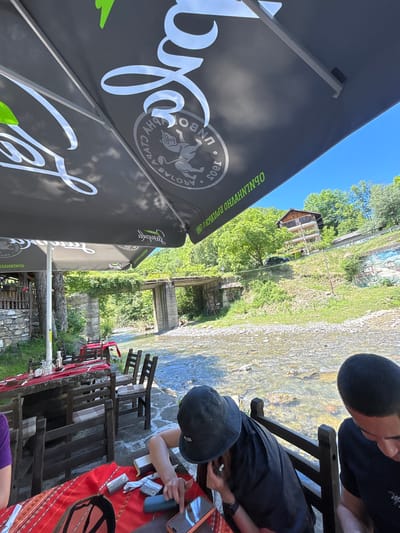 The image captures an outdoor dining area by a river in Beli Osam, Bulgaria. Two individuals are partially visible at a table covered with a red tablecloth. One person, wearing a black cap, is seated and appears to be interacting with a mobile phone and possibly an earphone. The other person, only their head is visible, is also seated at the table.
The table is set with glasses and bottles. Wooden chairs surround the table. In the background, a shallow river flows over stones. A concrete bridge spans the river further upstream, and beyond that, a hillside is covered with lush green trees. A house is visible on the slope. The scene is shaded by a large black umbrella with white lettering and logos. The overall impression is of a relaxed outdoor setting, likely a restaurant or cafe, situated in a natural environment with a focus on the river and greenery.