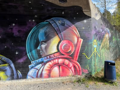 The image shows a concrete wall in Lørenskog, Norway, covered in graffiti. The primary focus is a mural of an astronaut with a helmet, depicted in a semi-realistic style with vibrant colors, set against a backdrop resembling a nebula or outer space with stars. Adjacent to the astronaut is a depiction of a tall, thin green alien figure with yellow markings. In the foreground, there's a dark blue trash bin standing on grassy terrain. The ground is gravel, and there is sparse vegetation along the base of the wall. Overhead, part of a concrete structure is visible, suggesting a bridge or overpass. Trees are visible in the background.