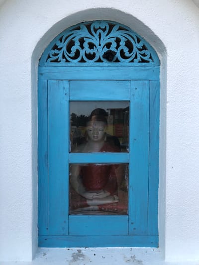 An arched, bright blue wooden window is set into a textured white wall. The window has a glass pane divided into two sections. In the upper section, decorative wooden filigree is visible. Through the glass, a statue of a Buddha is seated in meditation, wearing a red robe. The statue is illuminated and appears to be within a structure behind the window. The lower section of the window also shows the Buddha's legs and feet. The white wall is roughly plastered and shows some signs of wear and dirt at the base. The overall scene is simple and focuses on the architectural elements and the religious statue. The image is likely taken outdoors in Pilana, Sri Lanka, given the architectural style and the presence of a Buddha statue.