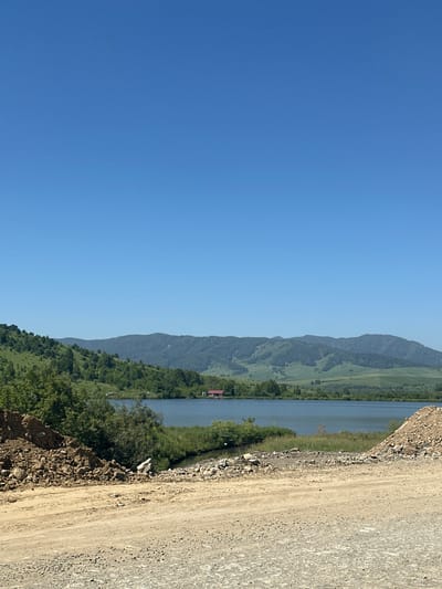 The image depicts a serene natural landscape under a clear blue sky. In the foreground, there is a dirt and gravel road, suggesting a rural or undeveloped area. To the left, a pile of excavated earth and rocks is visible, hinting at recent construction or land management activities.

Beyond the foreground, a tranquil lake stretches across the midground, reflecting the sky. The lake is bordered by lush green vegetation, including trees and reeds. On the far side of the lake, rolling hills covered in dense green forests rise up, forming a picturesque backdrop. In the distance, larger, more subdued mountain ranges complete the scene. A small red-roofed building is nestled amongst the trees on the northern shore of the lake, adding a touch of human presence to the otherwise natural environment.

While the image does not explicitly show the city of Altaiskoe, Russia, the landscape is consistent with the geographical features of the Altai region, which includes many lakes, forests, and mountains. The overall impression is one of natural beauty and peacefulness, perhaps a location outside of a developed urban center.