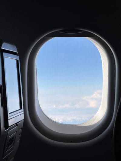 The image shows an interior view through a rounded window, likely from an aircraft or spacecraft. Through the window, a blue sky with light clouds is visible. Below the window is a dark-colored control panel or device with a small rectangular screen, reflecting the sky. The surrounding interior is dark and appears to be made of a smooth, possibly molded material. The location, Stolbishche, Russia, is not discernible from the image.