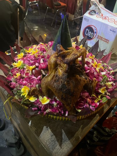 A whole roasted chicken is presented on a platter decorated with colorful flower petals, specifically pink and yellow ones, and dark red leaves. A green conical centerpiece, likely made of banana leaves, is positioned behind the chicken. To the right of the platter, a white box with red and blue branding that reads "Cleo" and "Tanubel" is visible, along with a striped object and red plastic bags on the floor. The platter rests on a wooden surface. In the background, out-of-focus individuals are seated at tables with chairs. The scene appears to be set indoors.