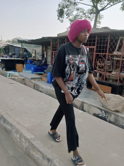 An outdoor urban scene in Mumbwa, Zambia, depicts a young person walking on a concrete pathway. The individual, positioned in the foreground and center-right, has bright pink hair and wears a black oversized t-shirt with a graphic print featuring two figures, one identifiable as Tupac Shakur. They are dressed in black trousers and black two-strap sandals with white soles. A light-colored strap is visible over their right shoulder. Their right hand is slightly raised. To the right, a large, light-colored rock rests at the edge of the pathway. The background consists of a street lined with structures. On the right, a building with a brown corrugated roof and decorative metal security grates on its windows is visible, with a tree displaying bare branches above it. To the left, an informal market or residential area contains several individuals, primarily men. At least two men are seated on blue upholstered sofas in an open-air setup. Other men are standing amidst various items including blue containers, mattress cushions, and tables. The sky is pale and overcast.