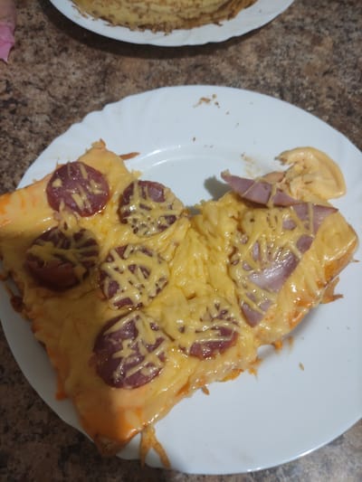 A close-up, high-angle shot shows two slices of pizza on a white plate. The pizza is covered in melted cheese and has toppings of sliced pepperoni and pieces of ham. The background is a textured brown countertop. The image is located in Kobeliaky, Ukraine.