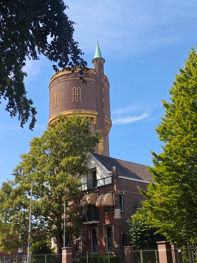 The image shows the Water Tower of Tilburg, Netherlands, a historic brick structure with a conical green roof. The tower is cylindrical with a decorative yellow band and arched windows. In front of the tower is a lower brick building with a tiled roof and striped awnings over the windows. Lush green trees frame the water tower, with one large tree in the foreground obscuring some of the lower building. A streetlamp and a fence are visible in the lower portion of the image. The sky is a clear blue with a few wispy clouds.