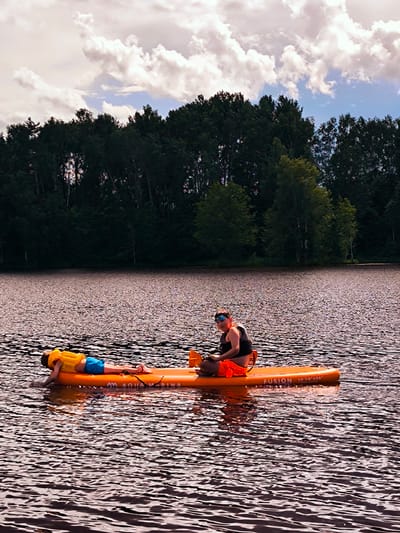 Two people are on an orange paddleboard on a body of water, likely a lake, with a line of dark green trees along the far bank under a sky with scattered white clouds. In the foreground, the water has a rippled surface with dark reflections. On the paddleboard, a person wearing a yellow life vest and blue shorts is lying face down. To their right, a person wearing a black top, orange shorts, and sunglasses is sitting and holding a paddle. The paddleboard has "AQUA MARINA" and "FUSION 10'10"x32"" printed on it. This scene is consistent with being at the Daugava River near Vecumnieki, Latvia, a popular spot for watersports.
