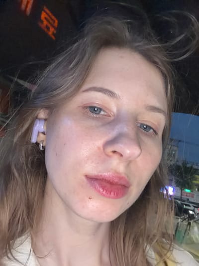 A close-up selfie shows a young Caucasian woman with light brown hair and blue eyes. She is wearing a pale yellow top and a light-colored garment underneath. A single, light purple earbud is visible in her left ear, and a small hoop earring adorns her left earlobe. Her skin appears fair with some freckles and a slight sheen, and she is wearing light pink lipstick. The background is blurred, but suggests an urban setting at night, possibly Uijeongbu-si, South Korea, with neon lights and the outline of buildings visible. The image is taken from a slightly high angle, looking down at the subject.
