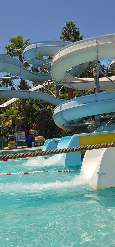 The image captures a bright, sunny day at a water park, likely in Algeciras, Spain, given the context. The main subjects are the prominent blue and white water slides, curving and twisting against a clear, vibrant blue sky. Lush green palm trees and other foliage surround the base of the slides, adding a tropical feel to the scene.

The activity taking place is the operation of the water slides, with water actively flowing down one of them, creating a splash as it enters the turquoise pool. A thick, braided rope with red and white floats marks a lane in the pool, further indicating a recreational water setting. The water itself is clear and reflects the sunlight, creating shimmering patterns on the surface.

Notable details include the scale of the water slides, suggesting a large amusement facility. The overhead perspective from which the photo is taken, along with the clear sky and bright water, points to a daytime scene, likely midday or early afternoon, given the intensity of the sunlight. The presence of greenery implies a warm climate suitable for a water park. While no people are clearly visible and identifiable in the image, the structure of the slides and the pool suggests people are present or expected to be present enjoying the facilities. No visible text is present in the image.