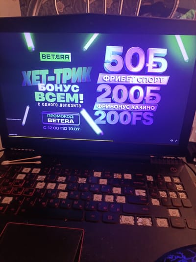 The image shows a laptop screen displaying an advertisement for "BETERA" which features a "HET-TRIK BONUS VSEM!" and offers "PRIBEТ SPORT 200B" and "FRIBONUS KAZINO 200FS". The advertisement also mentions a "PROMO CODE: BETERA" and a validity period from "12.06 TO 19.07". The laptop is placed on a surface, and the keyboard is partially visible, with several keys missing. Two cotton swabs are resting on the keyboard. The background of the image shows a blurred, patterned surface. There are no people visible in the image. The location of Александрыйскі сельскі Савет, Belarus is not depicted or suggested in this image.
