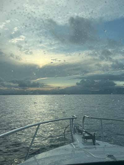 The scene presents a marine view from the bow of a white vessel, likely a motor yacht, situated on the waters offshore of Port Kembla, Australia, looking towards the distant coastline. The immediate foreground displays the boat's white foredeck, equipped with stainless steel bow rails and an integrated anchor roller system. The viewing surface, possibly a window or camera lens, is covered with numerous small, distinct water droplets. The midground consists of a vast expanse of dark, rippled water, reflecting the overcast sky. In the background, a low-lying landmass stretches across the horizon, consistent with the Port Kembla shoreline, with subtle elevated terrain or structures visible in the distance. A single, small, dark vessel is discernible on the water in the middle-right portion of the background. The sky above is largely overcast with stratified grey and blue clouds, featuring a prominent, diffuse yellowish-white light source on the left horizon, indicating either sunrise or sunset. No individuals or direct interactions are depicted.