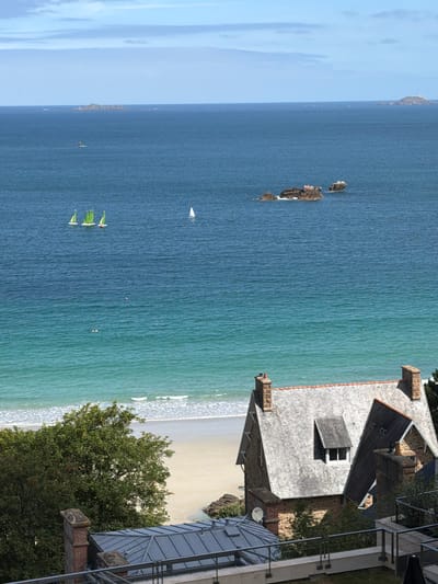 The image displays a picturesque coastal scene in La Clarté, France. The foreground features a stone house with a grey slate roof and a terra cotta chimney. A metal railing and a section of a modern balcony or extension are visible to the right of the house. Lush green trees grow on the grounds surrounding the house.
Beyond the house, the turquoise waters of the ocean meet a sandy beach, where gentle waves break. Several small sailboats with green sails are navigating the water. In the distance, a cluster of rocks emerges from the sea, and further out, a hazy outline of land can be seen. The sky is a pale blue with hints of white clouds. The lighting suggests it is daytime, with clear, pleasant weather. No people are explicitly visible, but the presence of the sailboats implies recreational activity on the water. The overall impression is one of a tranquil, scenic coastal setting.
