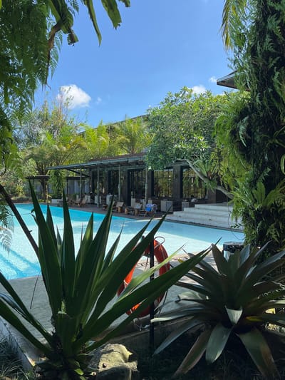 The image captures an outdoor swimming pool area in Petulu, Indonesia, surrounded by lush tropical vegetation. The pool is filled with clear, light blue water, suggesting a sunny day. A bright orange life preserver is propped up next to the pool edge. A covered structure with dark frames and a gray roof runs parallel to the pool's length, furnished with chairs and tables, some of which are occupied by people. The sky is visible between the foliage, showing a clear blue with a few scattered white clouds. The scene is framed by dense greenery, including large ferns and other tropical plants.