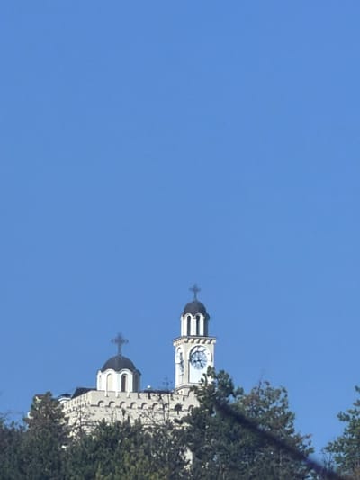 The image shows a clock tower and a church dome on a hill, partially obscured by trees. The sky is clear and blue. The clock tower is tall and white, with a round clock face and black hands. The church dome is dark and rounded, with a cross on top. The building below the dome appears to be made of stone. In the foreground, lush green trees are visible, suggesting a natural setting. The location is the city of Hadzhidimovo, Bulgaria.