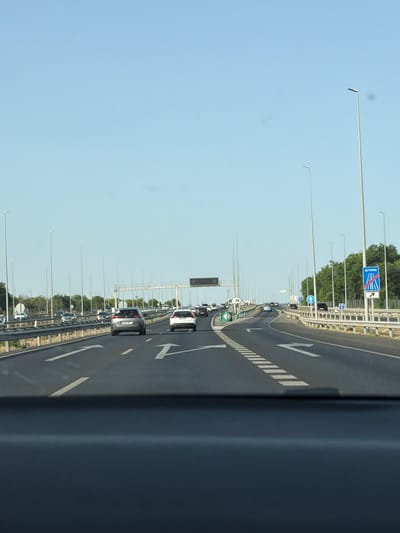The image depicts a clear, sunny day on a multi-lane highway. The perspective is from inside a vehicle, looking forward. The highway has white lane markings, including directional arrows. Several cars are visible on the road, moving in the same direction. Streetlights are spaced along the side of the highway. There is a green traffic island with some signage on the left side of the road. On the right side of the road, a blue and white road sign indicates "AUTOVIA" with an arrow pointing to the right, suggesting an exit or a change in route. Trees and vegetation line the sides of the highway in the distance. The sky is a clear blue. The scene is consistent with a highway in a developed area, and the "AUTOVIA" sign specifically identifies it as a Spanish highway. There is no explicit information in the image to pinpoint the exact location of Las Nieves, Spain, but the "AUTOVIA" sign is a strong indicator of Spain.