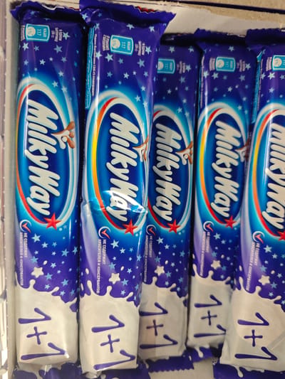 The image displays a close-up shot of several 'Milky Way' chocolate bars, positioned side-by-side in what appears to be a retail setting. The packaging is predominantly blue with white lettering and graphics, including the brand's logo and a '1+1' promotional offer. Nutritional information is printed on the packaging in Russian, including a calorie count of 117 kcal per serving. The chocolate bars are arranged in a row, suggesting they are stocked on a shelf or in a display case. The scene is brightly lit, highlighting the details of the packaging.