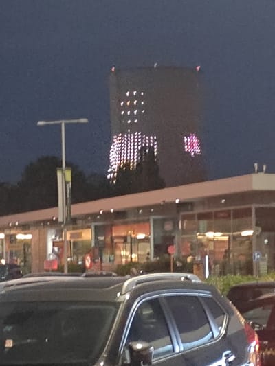 In Sint-Pieters-Leeuw, Belgium, at night, a tall building displays a pattern of illuminated pink hearts and white dots. In the foreground, the dark roof and side of a black SUV are visible, with the vehicle partially obscuring a low-rise building with illuminated windows. A streetlamp with a banner stands to the left of the SUV.