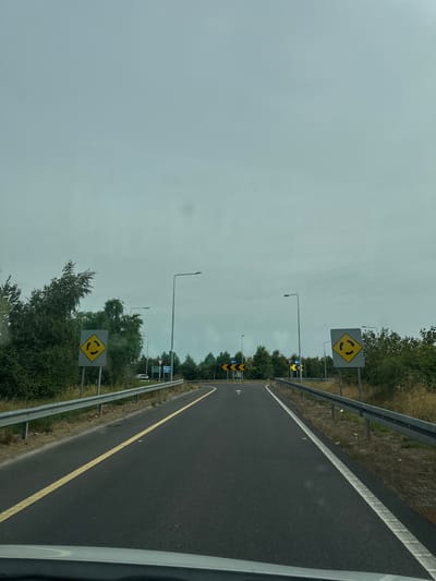 This is a first-person perspective, eye-level, outdoor, daytime, medium shot of a road in Father Campion Place, Ireland. The road is asphalt and has a yellow line on the left and a white line on the right. There are guardrails on both sides of the road. On the left side of the road, there is a yellow diamond-shaped sign with black arrows indicating a roundabout. Further down the road, there are more signs, including directional signs. On the right side of the road, there is another yellow diamond-shaped sign indicating a roundabout. The sky is overcast and appears light blue-gray. Trees and vegetation are visible on the sides of the road. The scene appears to be a highway or major road leading towards a roundabout.