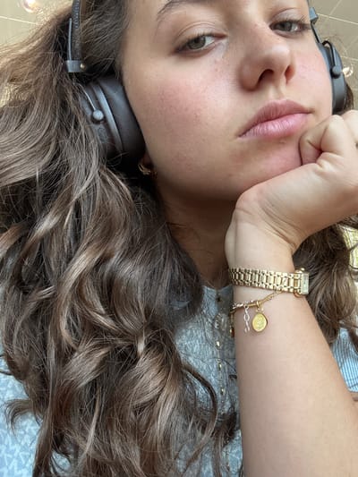 The image is a close-up, eye-level shot of a young woman's face and upper body. She is wearing black over-ear headphones. Her right hand is propped up, supporting her chin, and her left hand is visible on her wrist, adorned with a gold watch and a charm bracelet. The bracelet has a circular charm with text that appears to read "FAITH ON MARBLE." She has long, wavy brown hair. The background is out of focus, showing what might be a ceiling light and a patterned fabric. The overall lighting is natural and soft. The location is specified as Azzano San Paolo, Italy, but this is not visually evident from the image itself.