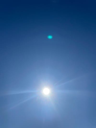 The image depicts a bright, clear blue sky with the sun visible in the upper right portion of the frame. A small, out-of-focus, light blue/green circular artifact is visible in the left portion of the frame. The scene appears to be a simple view of the sky, without any identifiable features to connect it to Поедуги, Russia or any other specific location. No people, subjects, objects, actions or interactions are present in the scene, beyond the celestial and optical phenomena.