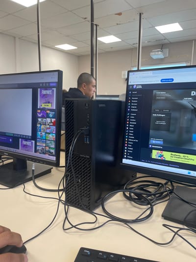 An indoor scene depicts a computer lab or classroom setting in France, consistent with a facility that could be found in Hérouville-Saint-Clair. The foreground features a light-colored desk surface with a black computer mouse held by a left hand and a partially visible black keyboard displaying an "Echap" key. Numerous tangled black cables are present on the desk. A black Dell OptiPlex 3080 SFF desktop computer tower sits centrally, with two black monitors on either side. The left monitor displays a web browser featuring "FORTNITE" advertisements and various game thumbnails. The right monitor shows the "CrazyGames.fr" website with categories such as "Action" and "Aventure," highlighting "DEADSHOT.IO" and an advertisement for "Creative Cloud Pro." In the mid-ground, a male individual with short, dark hair is seated at a workstation, facing right and concentrating on a screen. The background consists of plain white walls, a white ceiling with multiple square fluorescent light fixtures, several dark metal poles extending from floor to ceiling, and a white projector mounted on the ceiling to the right. Some minor surface damage is visible on ceiling panels.