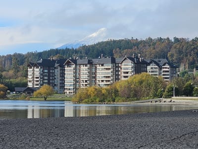 The image shows a view of Villarrica Lake in Villarrica, Chile. In the foreground is a black sand beach. The water is calm and reflects the buildings in the background. A row of apartment buildings is visible behind a line of trees, which show autumn foliage. Beyond the buildings, a snow-capped mountain rises in the distance, partially obscured by clouds. A person is sitting on a bench along the shoreline. The sky is overcast.