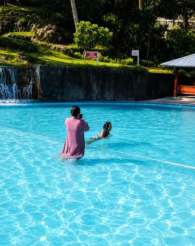 In a bright blue swimming pool, two individuals are visible. One person, facing away from the camera and wearing a long, flowing purple garment, stands partially submerged in the water, holding a mobile phone to take a photograph. To their right, a second person, a child, is swimming in the pool. The background features a lush, green hillside with trees and a waterfall cascading into the pool. Signs are posted on a stone wall above the pool, one red and white, and another white with black lettering. A wooden structure with a white roof is situated to the right, and a wooden bench is visible underneath it. The overall scene depicts a sunny day at a recreational pool. This image was captured at a resort or public swimming area in Silaiyajae, Indonesia.