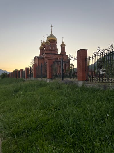 The image depicts an exterior view of a red brick building with golden domes, resembling a church or monastery. The building is partially visible along the right side of the frame, extending into the distance. It is surrounded by a decorative black metal fence and a brick wall. To the left of the fence is a lush green grassy area, with some taller grass blades and smaller plants. In the background, the sky transitions from a soft yellow near the horizon, suggesting it is either sunrise or sunset, to a pale gray at the top. Distant hills or mountains are faintly visible on the horizon. The overall atmosphere is peaceful and serene, enhanced by the soft lighting of the sky. There is no visible text in the image. The orientation of the image is vertical, meaning the subjects are rotated 90 degrees counter-clockwise from their natural orientation.