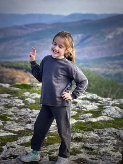 The image, originally rotated 90 degrees clockwise, depicts a young girl standing on a rocky surface outdoors, overlooking a vast mountainous landscape. The setting is identified as Родниковское сельское поселение, Ukraine.
The central subject is a smiling young girl with light brown hair. She is dressed in a dark grey long-sleeved sweatshirt and matching dark grey pants. On her feet, she wears light-colored sneakers with subtle blue and green details, and matching socks. She is posing for the camera, with one hand on her hip and the other slightly extended, and a wide, joyful smile on her face, indicating happiness and enjoyment of the moment.
The foreground consists of rugged, grey rock formations partially covered with vibrant green moss. Behind the girl, the background extends into a hazy mountainous or hilly terrain, displaying tones of blue and green, suggesting a significant distance. The sky above is bright but appears somewhat overcast or hazy, contributing to the soft, diffused lighting across the scene.
The time of day is daytime, likely late morning or early afternoon, characterized by the absence of harsh shadows. There is no visible text in the image. The scene captures a peaceful moment of a child exploring and enjoying a scenic, elevated natural environment.