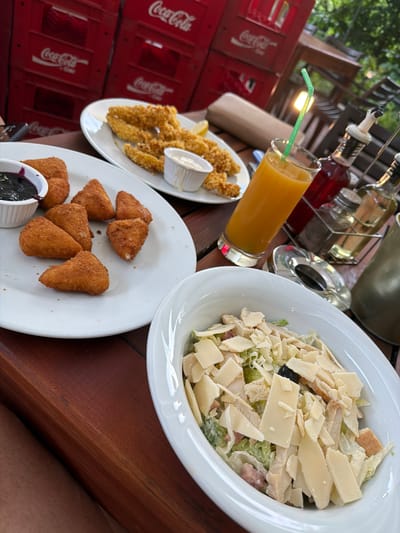 The image captures a meal setting on a wooden table. There's a Caesar salad in a white bowl, a plate of fried chicken with a small container of white sauce, a plate of fried cheese wedges with a small container of dark-colored sauce, and a glass of orange juice with a green straw. Condiment bottles and a metal ashtray are visible in the background. Coca-Cola crates are stacked to the left of the table. The specific location in Milanovo, Bulgaria is not discernible from the image. No people are fully visible, though part of someone's leg is visible in the bottom right corner. The focus is on the food and drink presented on the table.