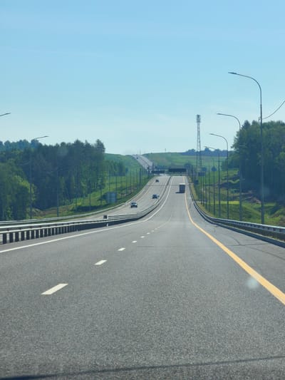 A low-angle, full shot captures a bright, sunny day on a multi-lane highway in Novaya Derevnya, Russia. The road stretches into the distance, with lanes marked by white dashes and a solid yellow line delineating the opposing traffic. Guardrails line the sides of the highway. On the left, a dense green forest covers a rolling hill. On the right, more trees are visible, interspersed with grassy slopes and a few streetlights. In the distance, a tall cellular tower stands against the clear blue sky. A few cars and a truck are visible on the road, moving away from the viewer. The scene is peaceful and depicts a typical day on a Russian highway.