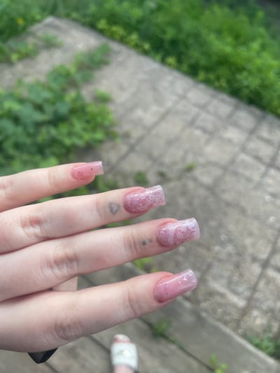 A close-up shot focuses on a person's left hand, adorned with meticulously manicured, square-shaped artificial nails. The nails are a light pink hue, with subtle glitter embedded within the polish. Two of the nails, specifically the ring and middle fingers, feature intricate white lace patterns applied over the pink base. Faint, dark grey heart-shaped tattoos are visible on the skin of these fingers.
The hand is positioned over a grey, paved surface that appears to be outdoors. The paving stones are laid in a grid pattern. Lush green foliage, some of it overgrown and wild, is visible in the background, suggesting a natural or semi-natural outdoor setting. The lighting suggests it is daytime, likely overcast given the soft illumination.
The location of Линёвка, Russia is not discernible from the provided image alone.