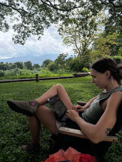 A woman is seated outdoors in a chair, looking down at a tablet device. She is wearing a sleeveless olive green top and dark shorts, with brown high-top Converse sneakers. Her legs are crossed, with one foot resting on a low wooden railing. The tablet is positioned on her lap, and her hands are on the keyboard. She has dark hair pulled back in a ponytail and a silver necklace. In the background, there is a lush green lawn, a wooden pathway, and dense foliage. Beyond that, a range of mountains is visible under a cloudy sky. The setting appears to be a natural, possibly tropical, environment.