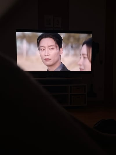 A man's leg is in the foreground, obscuring the lower left portion of the image. In the mid-ground, a television is turned on, displaying a scene from a show or movie. The screen shows two individuals. On the left is a man with dark hair styled forward, wearing a dark suit jacket over a grey shirt. He has a serious expression and is looking off to his left. To his right, a woman with dark hair is partially visible, her face turned away from the camera. The television is placed on a white media console with two open compartments, each containing a woven basket. Behind the television, a dark upright object, possibly a vacuum cleaner, is positioned near a wall. The room appears dimly lit, suggesting it is nighttime or the curtains are drawn. Two framed pictures are visible on the wall above the media console. The overall scene depicts a person watching television in a home setting. The location of Breisach am Rhein, Germany is not discernible from the visual information present in the image.