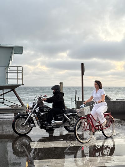 In Oceanside, California, a person rides a motorcycle and another person rides a red bicycle on a wet street with a crosswalk. The motorcycle is black and the rider is wearing a black helmet and jacket. The bicycle has a white basket with items inside and the rider is wearing white pants and a white polo shirt. In the background, there is a lifeguard tower, the ocean, and a cloudy sky. The scene appears to be near the coast.